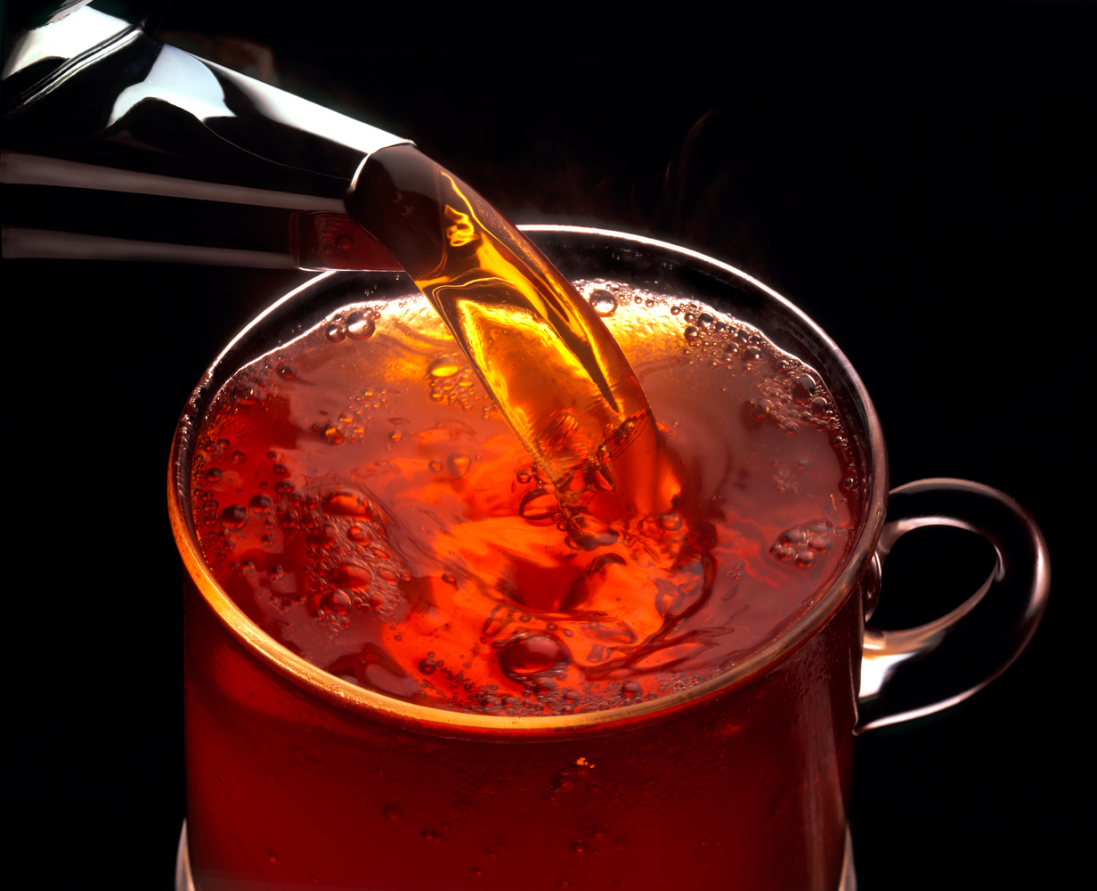 a shinny metal tea pout spout pours hot redish tea into a glass cup